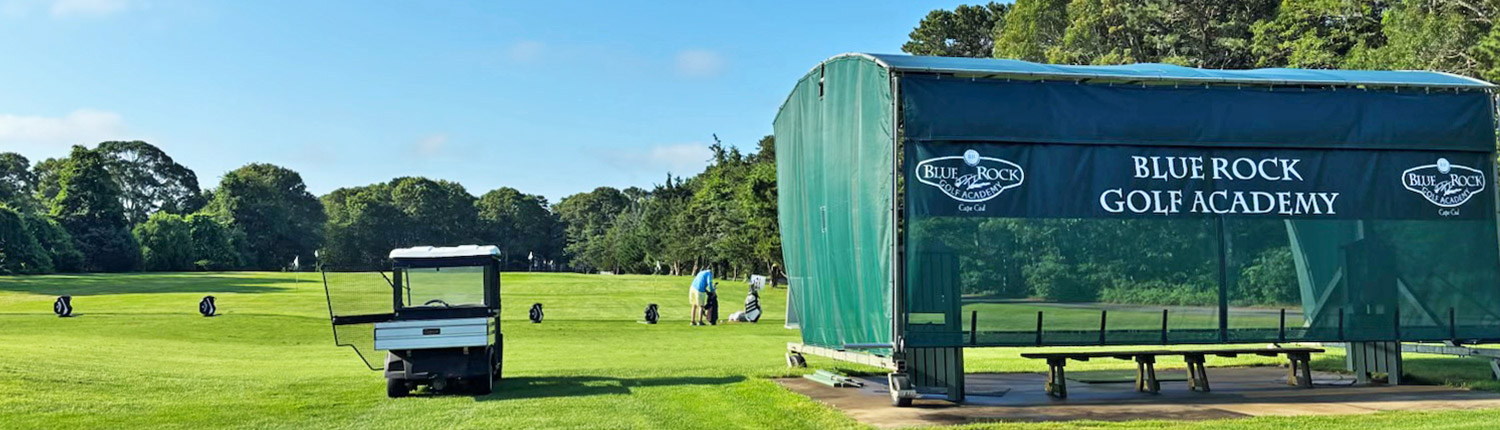 A golf practice range with a white golf cart, a person preparing to hit golf balls, and a covered practice area labeled “Blue Rock Golf Academy” on a sunny day.